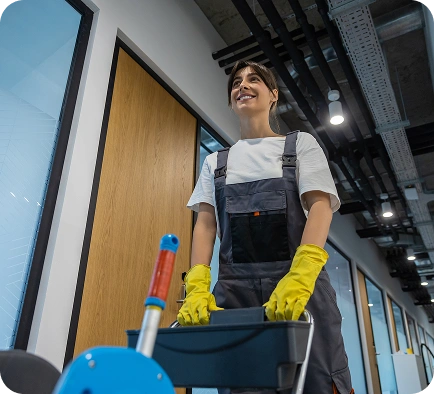 Uśmiechnięta osoba w kombinezonie i żółtych rękawiczkach pcha wózek sprzątający po nowoczesnym, dobrze oświetlonym korytarzu biurowym, sprawiając wrażenie profesjonalizmu i pozytywnego nastawienia