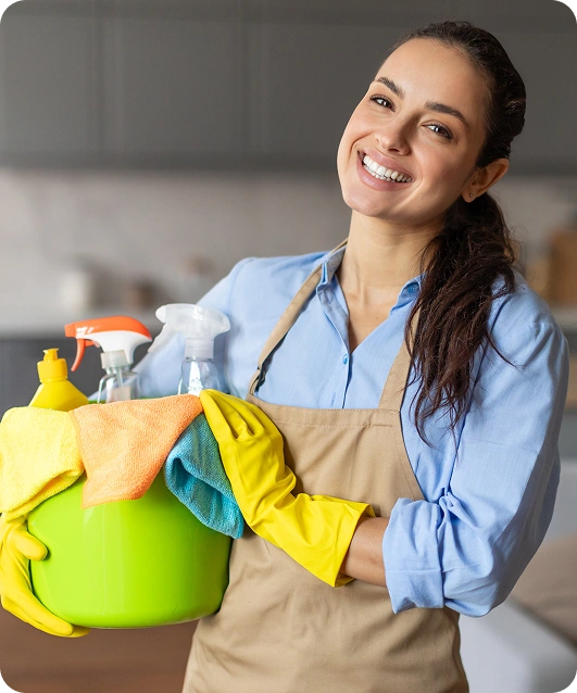 Uśmiechnięta kobieta w fartuchu i żółtych rękawiczkach trzyma zielone wiadro z środkami czystości w przytulnej kuchni. Scena ta oddaje czystość i pozytywne nastawienie
